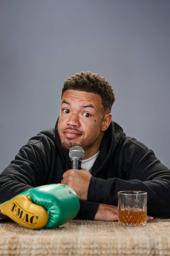 A man holds a microphone while sitting on a table, boxing gloves and glass of whiskey on table in front of him. He looks engaged and ready to entertain